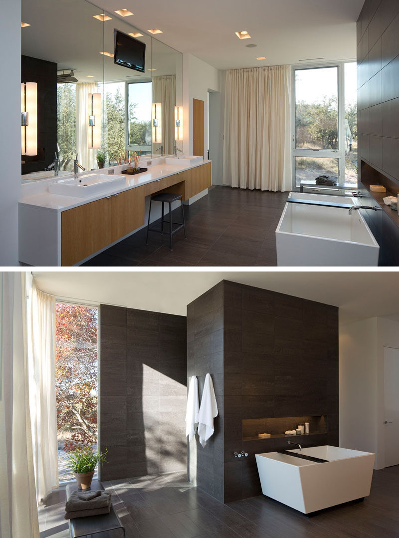 In this modern master bathroom, a floating double vanity with light wood cabinetry and white counters has a central cutout, just the right size for a stool and provides the perfect spot to sit and apply makeup. A stand alone tub is positioned against a partial wall covered with the same tiles as the floor. A built-in shelf in the wall next to the tub makes sure that everything you need while bathing is always within reach.