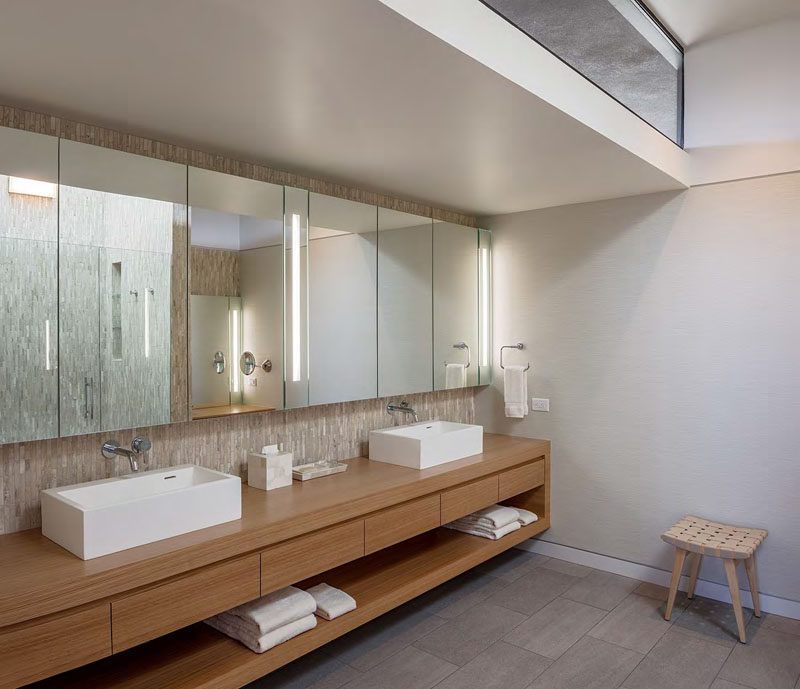 In this modern master bathroom, natural light from the clerestory window filters through to the space, while large mirrors fill the width of the wall. A floating wood double-sink vanity has plenty of drawer space and open storage.