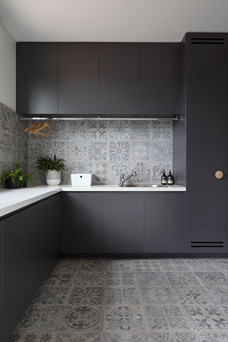 In this modern laundry room, matte black cabinets provide a strong contrast to the light grey tiles and the white countertops. A drying bar has been installed to hang drying clothes, and a sink makes clean-up or soaking clothes easy.