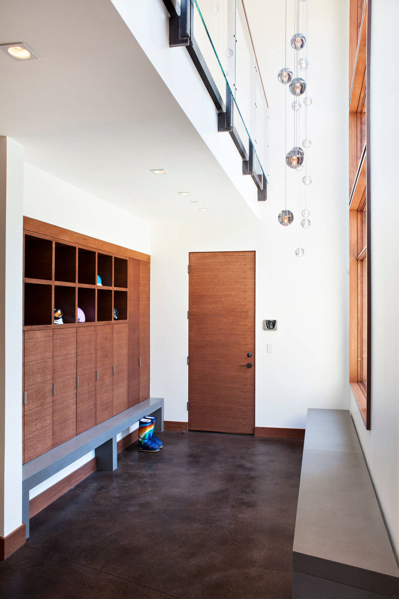 In this wood and white modern mudroom with concrete floors, built-in cabinets and lockers provide storage and organization for each family member, and multiple long concrete benches make it easy to put your shoes on. A wall of windows keeps the room bright, and a light feature hangs from the partial double-height ceiling. 