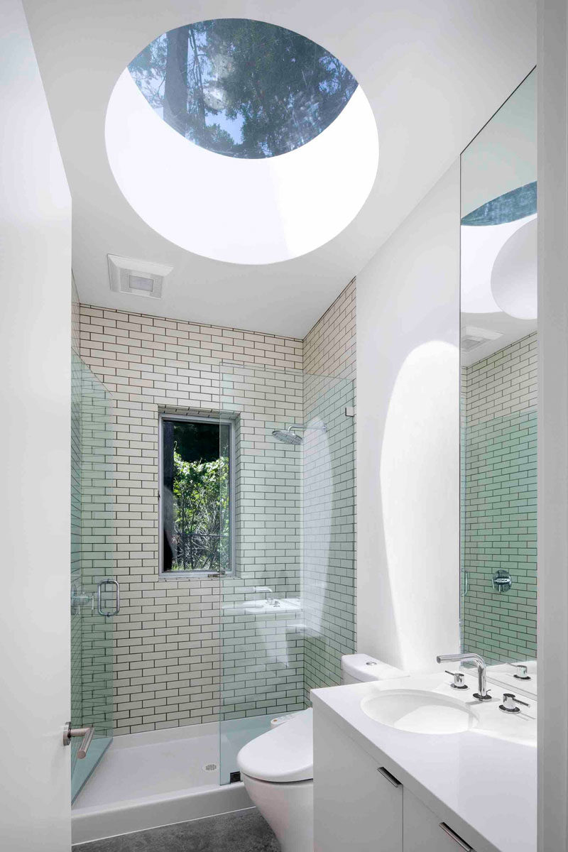 In this modern bathroom, a large circular skylight adds a lot of natural sunlight to the space, while the white walls and white vanity have been paired with a cream-colored rectangular tile to help reflect the light. A glass shower door keeps the rest of the room dry, and a small window lets you look out into the trees as you are showering.