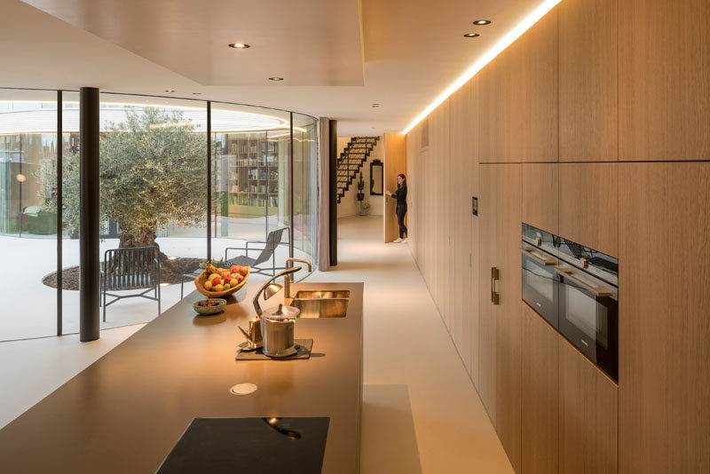 In this modern house, a wall of wood cabinets turns into the kitchen, with a large island. Hidden lighting at the top of the cabinets provides a soft glow to the space.