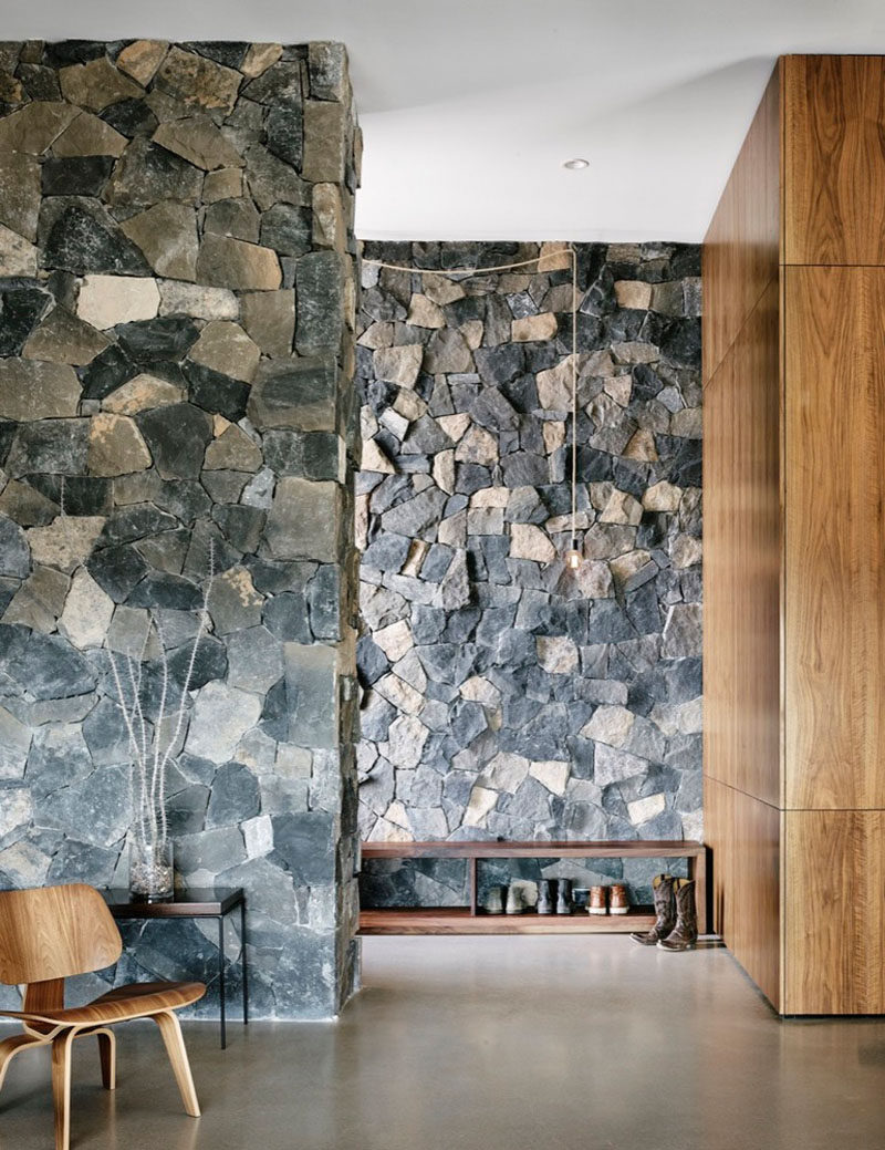 In the entryway of this modern house, grey stone lines the walls while American black walnut has been used for the wood cabinets. A wooden shoe rack helps keep the family shoe collection under control and an exposed hanging bulb brightens the hall and adds a rustic touch.