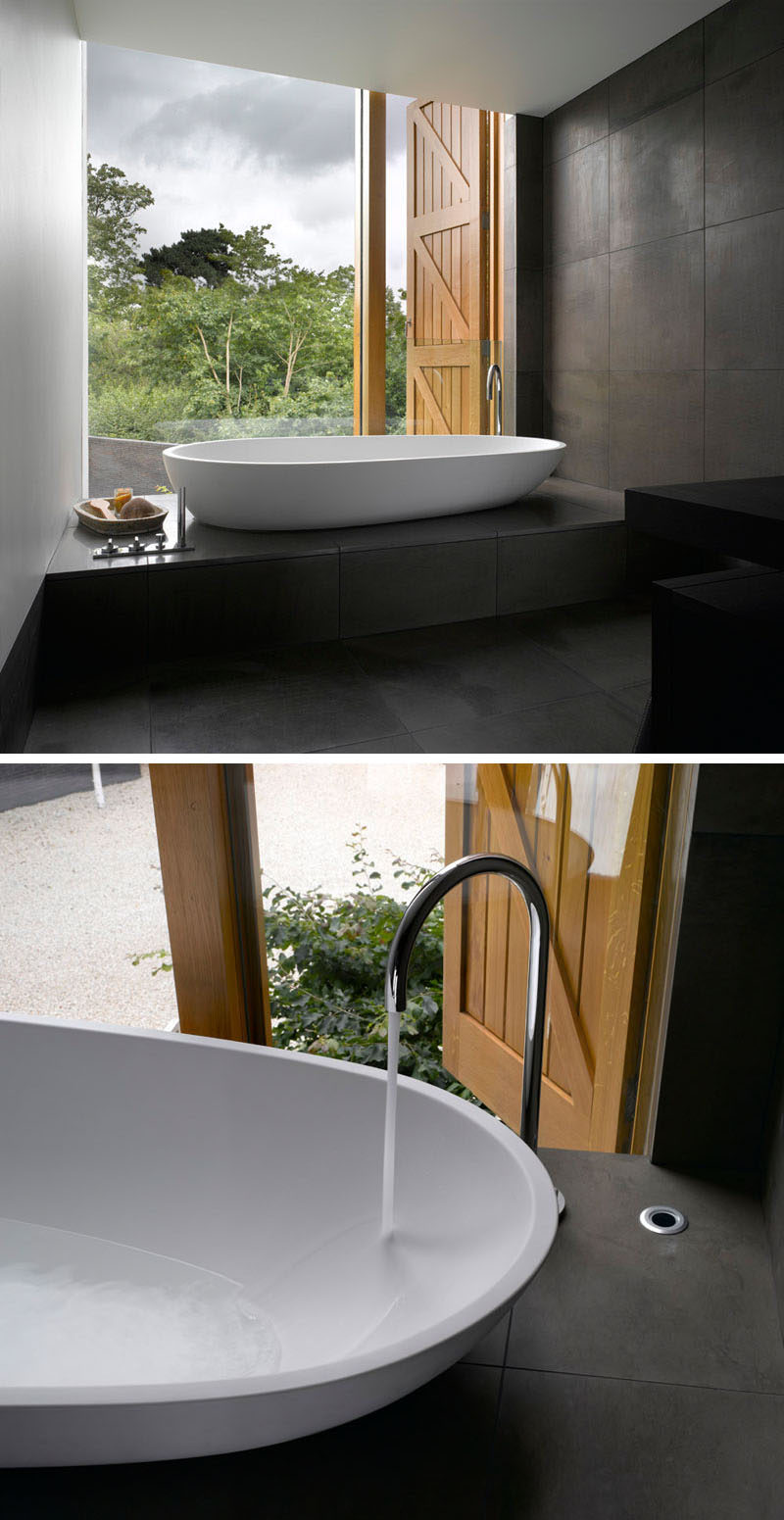 In this master bathroom, dark tiles have been paired with white walls and bathtub for a minimalist look, while the large windows provides views of the garden.