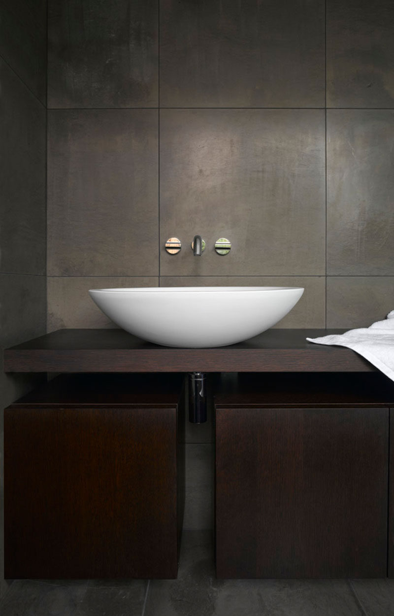 This dark and dramatic master bathroom features dark tiles, a dark wood vanity and a simple white basin.