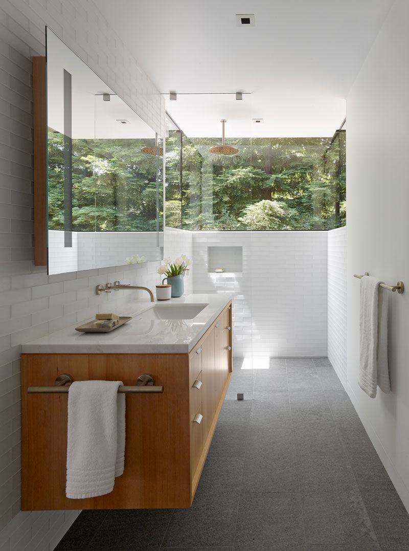 This modern and updated bathroom has white walls and white tiles, and a wrap-around window makes you feel like you are showering in nature. A light wood vanity with white countertop, light grey tile flooring and a large mirror help to keep the bathroom bright.