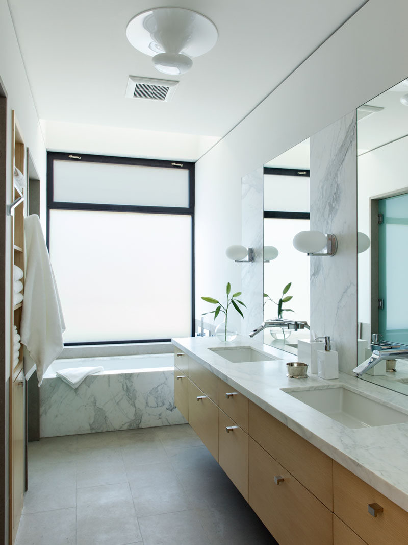 This modern bathroom features a floating light wood vanity with a stone counter and dual undermount sinks. A frosted window provides privacy while you're in the bath, but at the same time lets the light in to the room.