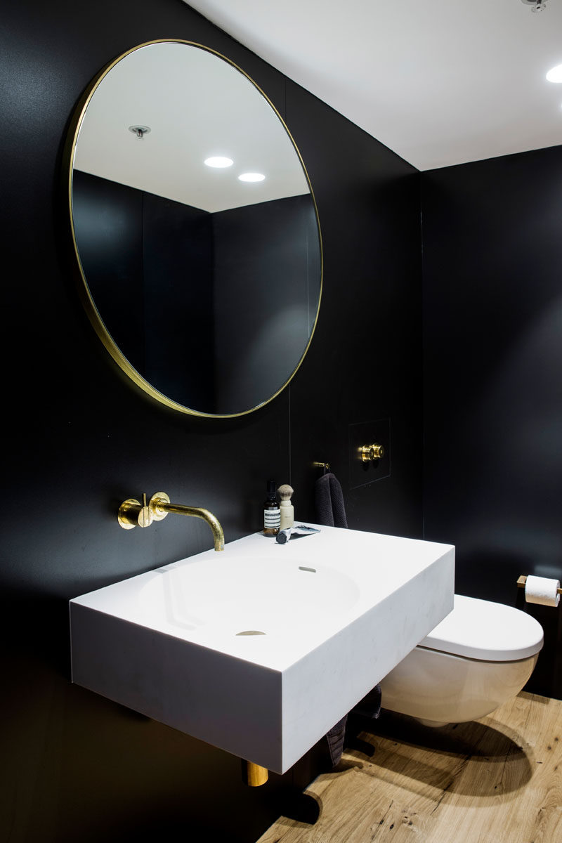 This modern and dramatic bathroom features black walls, a gold framed round mirror, a gold faucet and accents, a white vanity and wooden floors.