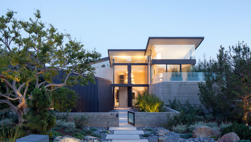 The front of this modern house has a path surrounded by landscaping, that leads to the large, light wood, pivoting front door.