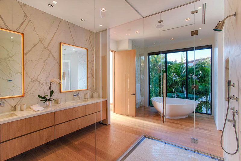 This modern master bath has a dual sink vanity, a shower with a floor-to-ceiling glass surround, and a white freestanding bathtub with views of the palm trees through the large windows.