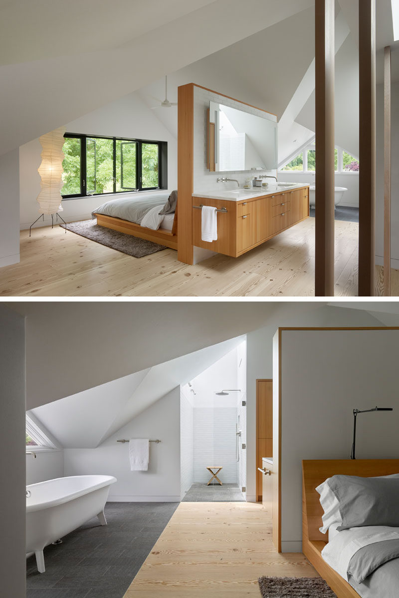 This modern master bedroom and bathroom suite look out to the treetops, while a tall white and wood headboard sits behind the bed and doubles as a wood vanity for the bathroom. Off to the side is a standalone bathtub and walk-in shower with white tiles and a glass shower door.