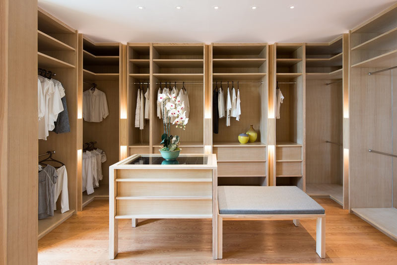 This large walk-in closet has plenty of light wood open shelving as well as drawers for storage. In the center of the closet is an accessories island and an upholstered bench.