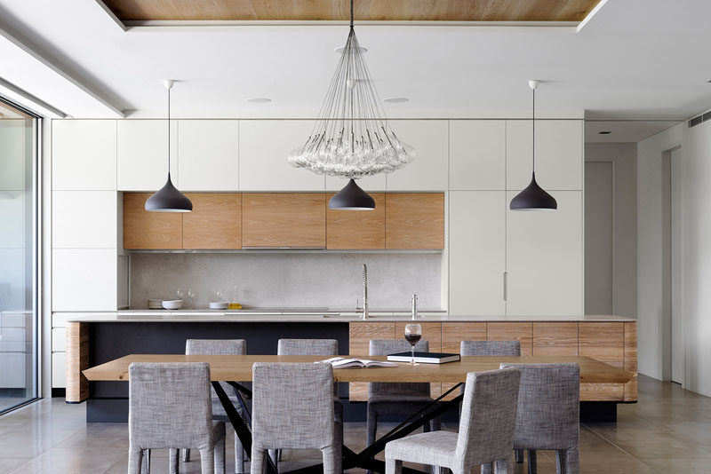 This modern kitchen features a stone countertop, solid timber doors and over-sized ceramic floor tiles. The dining room is made up of a large wood table, upholstered dining chairs and a large pendant lamp hanging above.