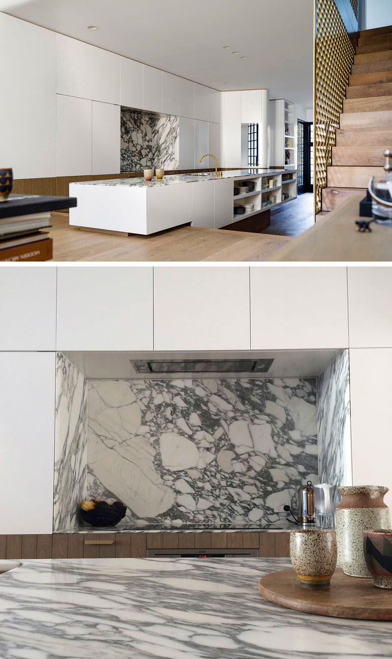 The living room in this modern house sits slightly higher than the kitchen, with the kitchen island straddling across both levels. In the kitchen, Carrara marble has been used for the countertop, and hardware free white and wood kitchen cabinets keep the space streamlined.