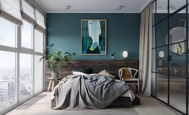 In this modern bedroom, a deep teal and wood accent wall provides the perfect backdrop for the artwork and bed, while black framed glass walls separate the bedroom from the living and dining area and allow the light from the windows to travel throughout the small apartment. At night, blinds and curtains can be drawn to provide privacy. #TealWall #ModernBedroom #WoodHeadboard #BedroomDesign