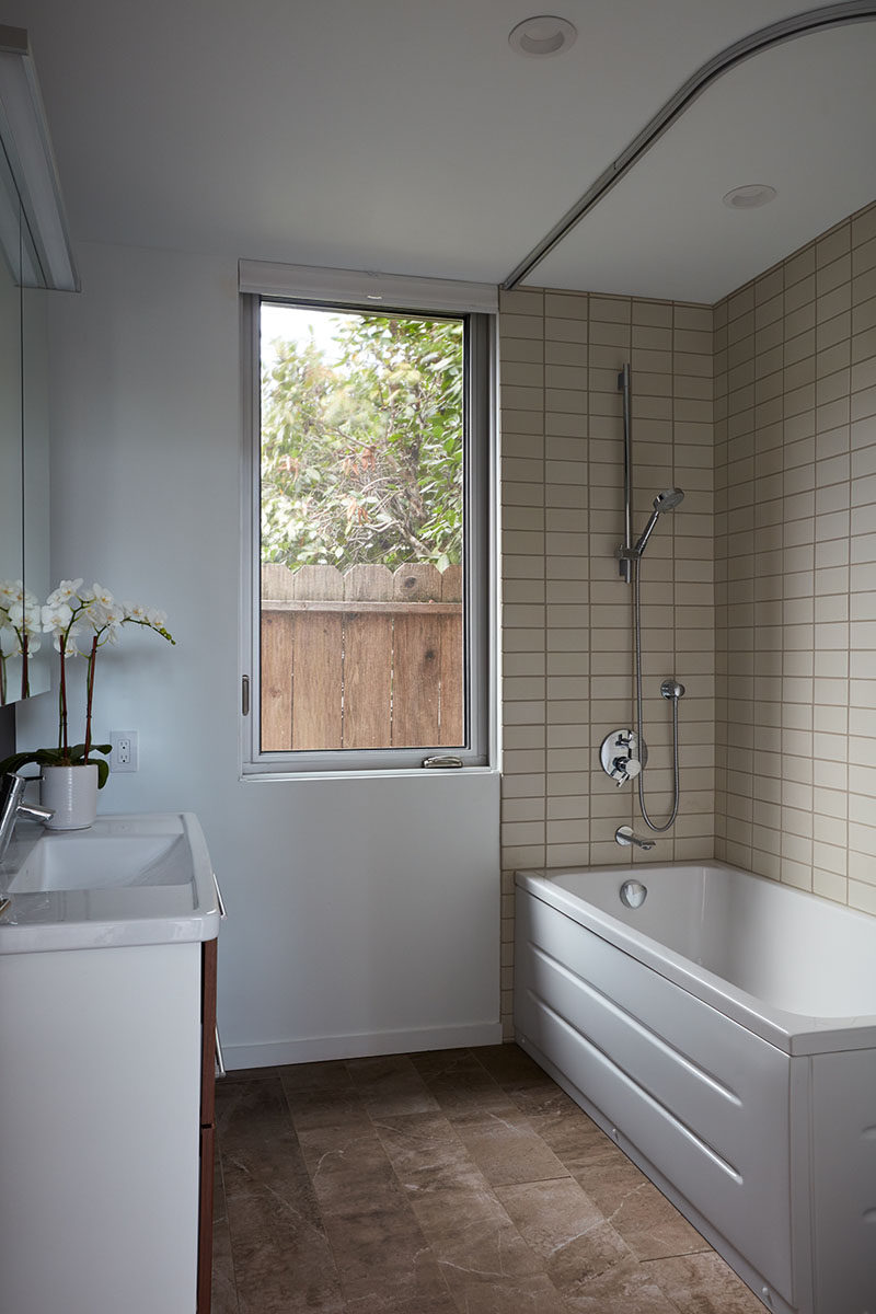 This simple bathroom has a tall window, and a large bath tub with shower is surrounded by cream colored rectangular tiles.