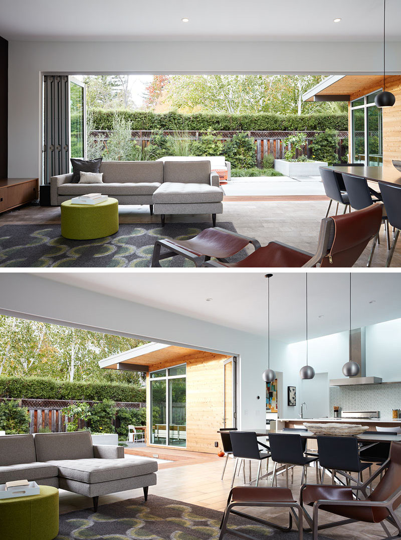 This modern open-plan living room looks out onto the patio through a wide, glass folding wall system. The gray, green, brown, and wood furniture match the colors of the outside space.