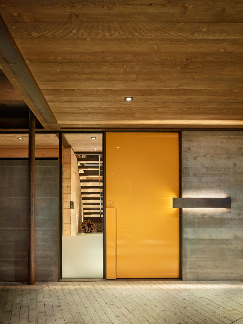 The vibrant yellow door of this industrious home is placed beside a window, giving a glimpse of the interior of this modern house.