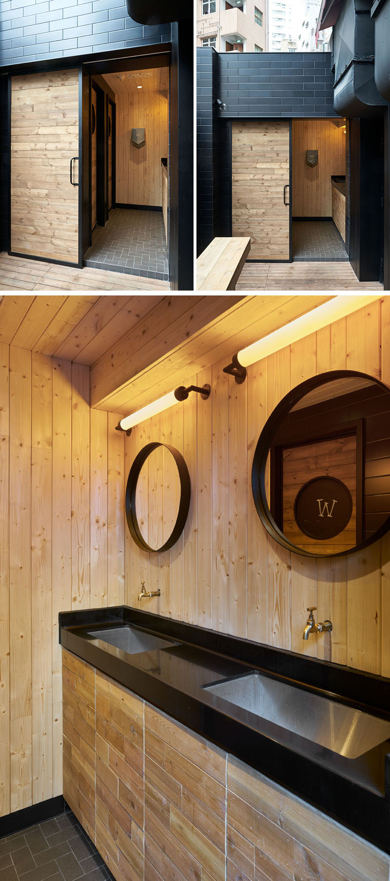 This modern coffee shop has a set of minimally decorated washrooms that stand out on a black wall with wood sliding doors. The interior is full of light wood, and the vanity features a black countertop with undermount sinks, and hanging above are two black framed circular mirrors.