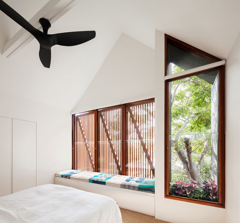 This mostly white modern bedroom has wood window frames and a built-in window seat with storage and an upholstered cushion. A small window box adds a touch of nature to the room.