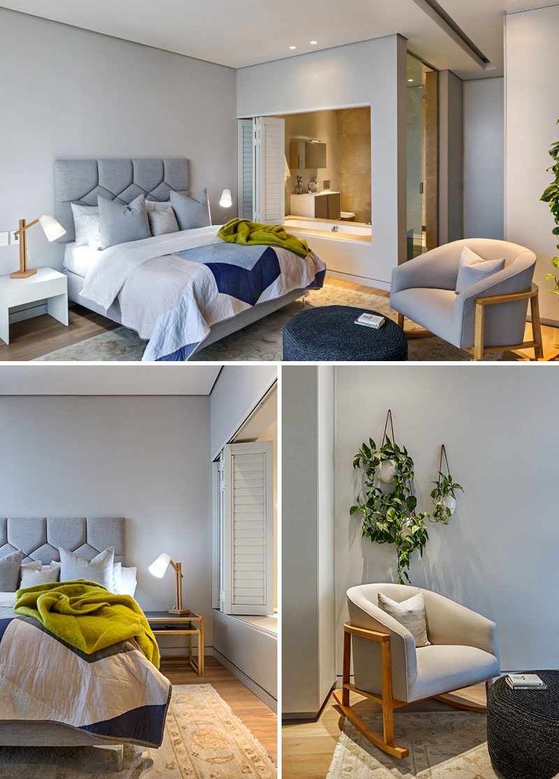 This modern bedroom is decorated in greys, blues, and whites, with a wood and grey upholstered chair and a large foot stool. There is an ensuite washroom behind a set of closing white shutters. 