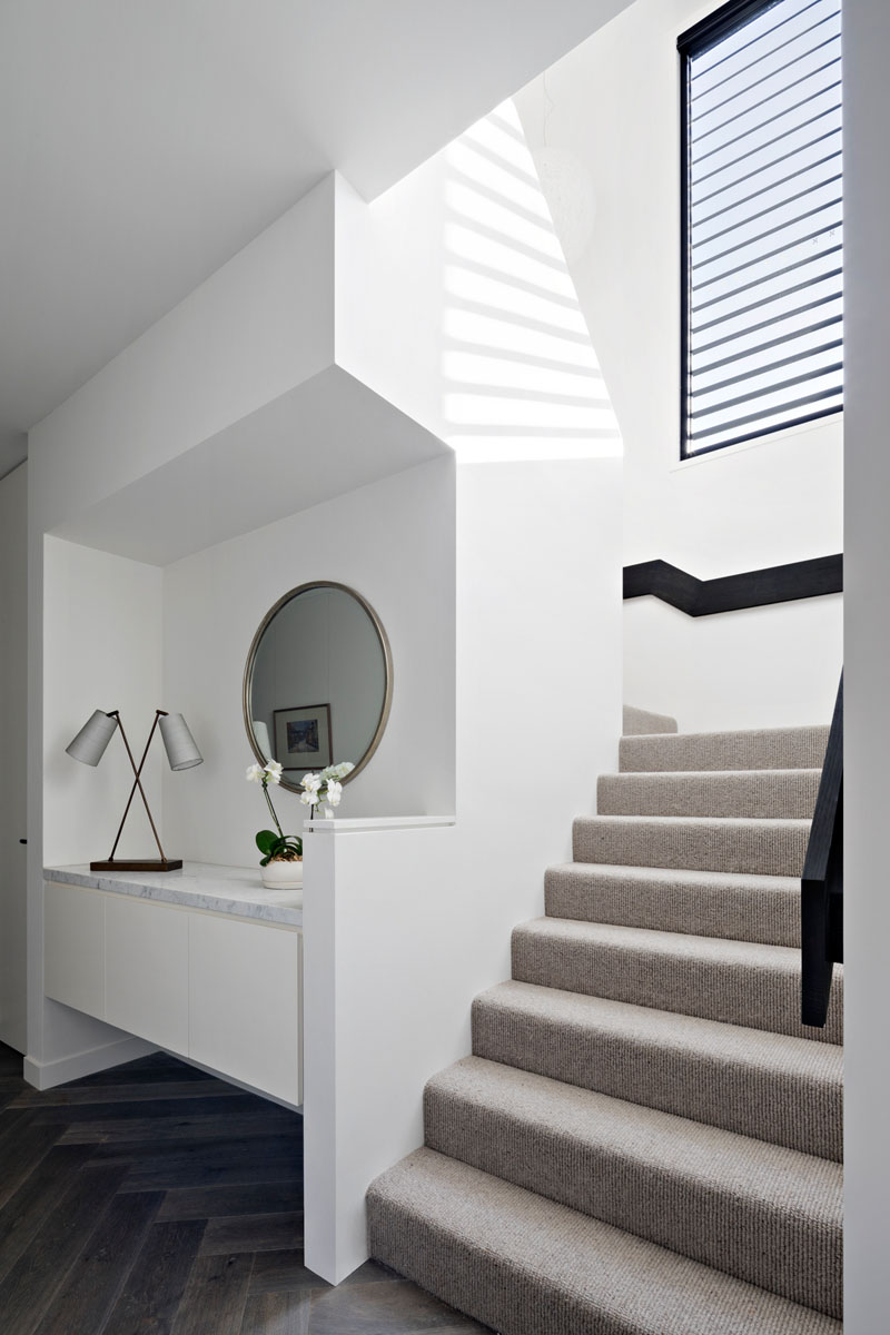 This modern hallway has a floating built-in cabinet, with a light colored carpeted staircase and a black railing that leads to the upper floor, with a matching black window frame.