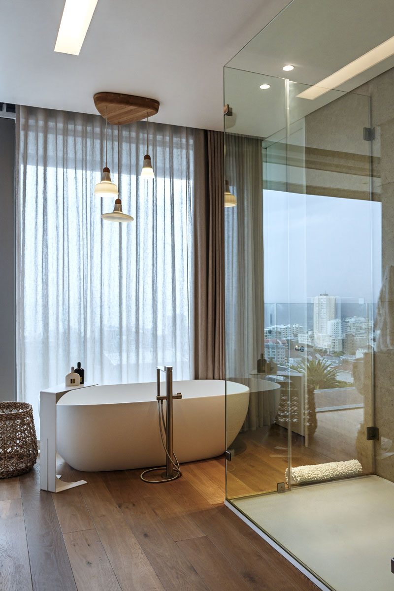 This modern master bathroom has a freestanding bath tub that sits beside large windows and a glass shower surround.