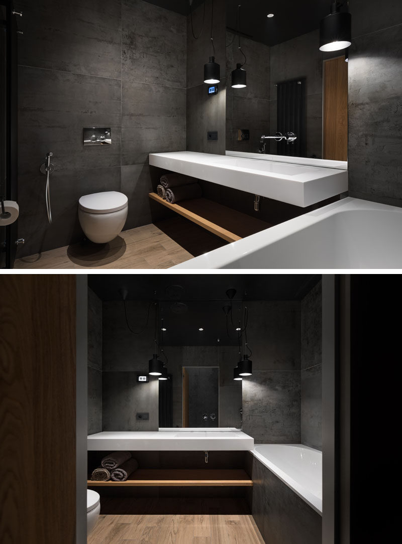 This bathroom is made up of concrete with wooden accents and flooring, and hanging light fixtures. The pure white floating countertop below the mirror has a seamless sink, while the matching bathtub has a surround that matches the walls.