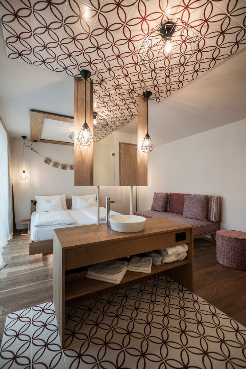 This modern hotel room has an open floor plan that's broken up by a wood vanity with a hanging mirror. The bathroom is defined by red and white patterned tiles that match the red sofa. 