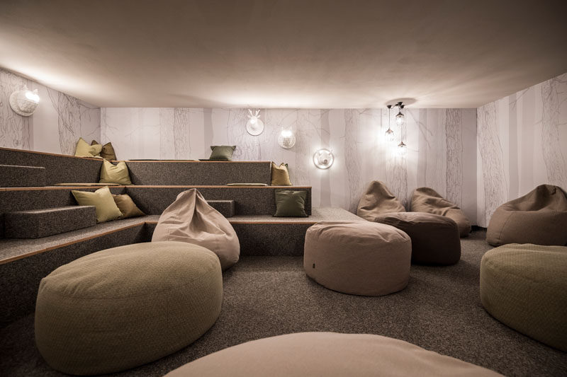 In this modern hotel, there's a relaxation room with floor to ceiling tree lined wallpaper, gray carpeted, and tiered benches. Animal light fixtures, and earth toned cushions and bean bags make this a perfect place to relax after adventuring in the wild.