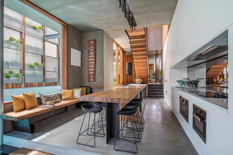 In this open kitchen, there's a wood bench with cushions, and opposite there's a wall full of white kitchen cabinets, as well as the oven and stove. A mirrored backsplash helps to reflect light in the room and make it appear larger.