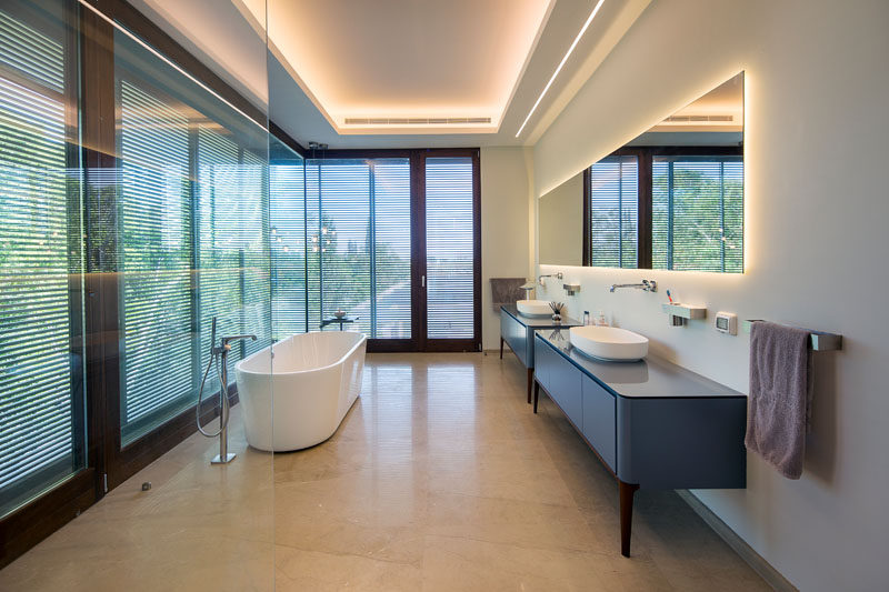 In this modern master bathroom, lighting is hidden from view in the ceiling and behind the large rectangular mirror. Double vanities provide plenty of space and a white freestanding bathtub sits next to floor-to-ceiling windows with privacy screens.