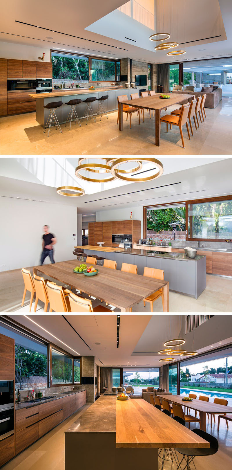 A void in the ceiling with circular pendant lights helps to anchor the dining table in the open plan room. Behind the dining area is the kitchen with a large grey island with a wood bar for sitting at. Wood cabinets line the wall and a large sliding window looks out onto the garden.