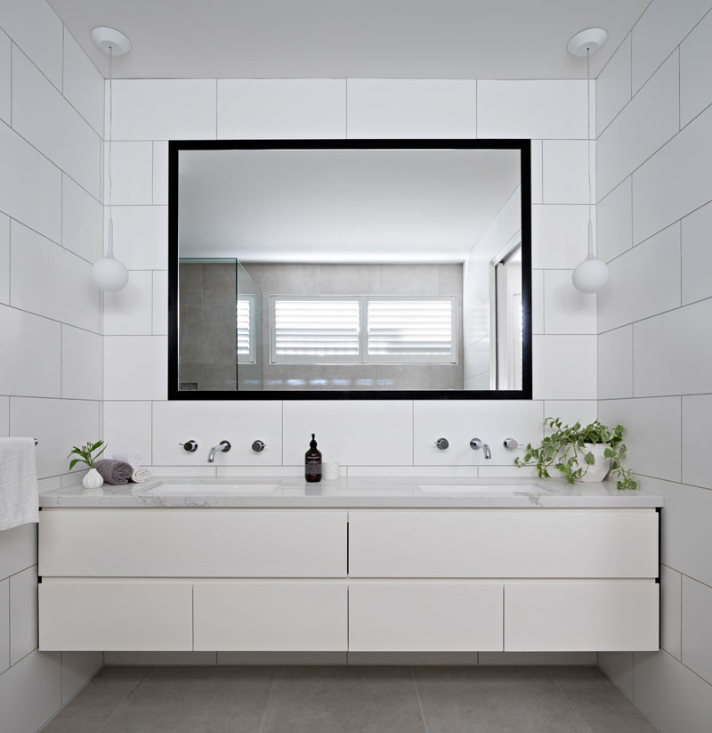 A large black framed mirror stand outs in this white modern bathroom that features large rectangular tiles on the walls . Two white pendant flank the mirror and hang above a marble counter with dual sinks.