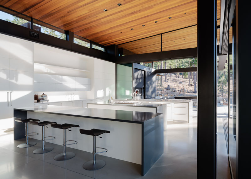 This modern kitchen that consists of three large white islands with grey countertops. The stained cedar ceiling stands out above the concrete floor, while white cabinetry sits below a set of clerestory windows, providing even more light to the open space. 