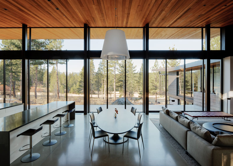 An oval dining table sits below a large white pendant light, and separates the kitchen from the living room. Floor-to-ceiling glass walls give an expansive view of the terrain outside while eating. 