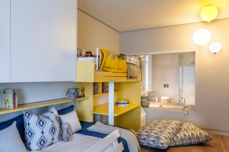 This modern kids bedroom is decorated in blue, white, yellow and balloon light fixtures. A white and yellow bunk bed has a desk below it, and a cut-out in the wall with shutters reveals the ensuite bathroom.