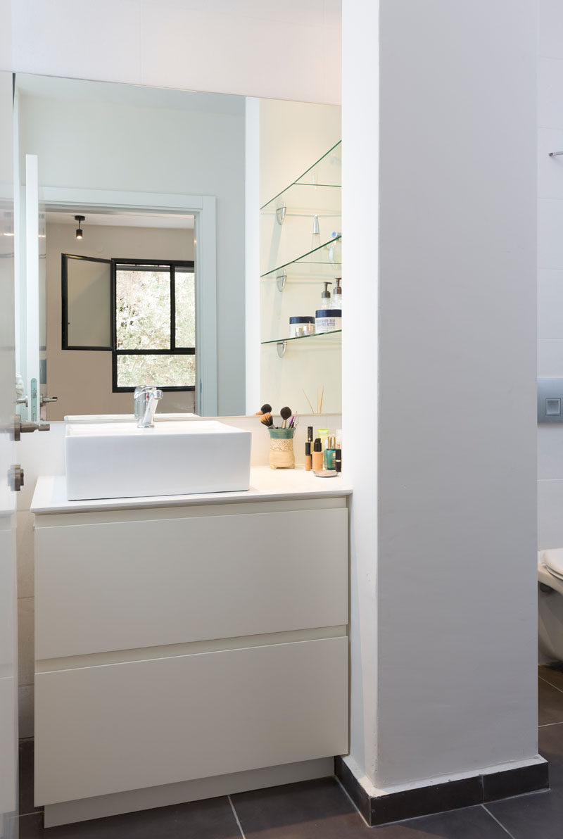 In this small modern bathroom, a white countertop sink with pull-out drawers is placed opposite the window. Accessible glass shelving has been placed to the right of the sink, separating the vanity from the toilet.