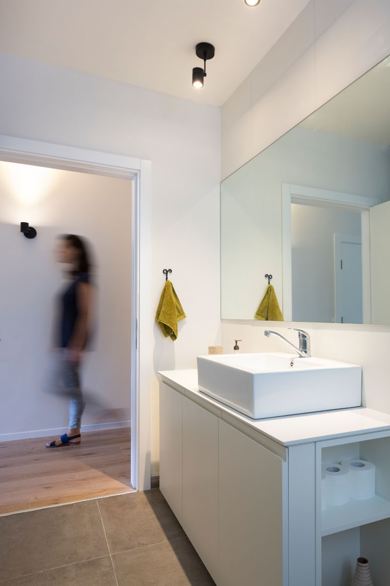 In this modern bathroom, a large mirror hangs above a square white sink and long vanity, while cabinetry below it has open shelves on one side. 