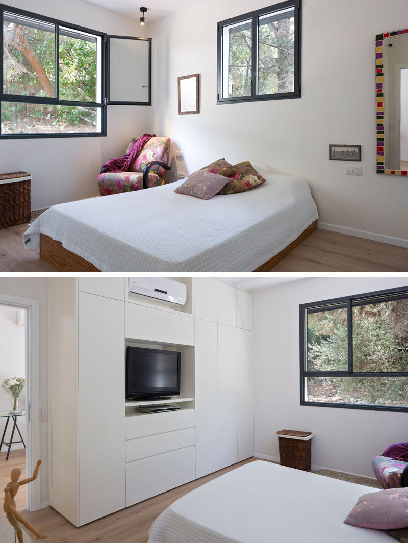 In this simple and mostly-white bedroom, black framed windows are used to emphasize the trees outside. White cabinetry blends in with the walls, making the space feel larger than it is. A pink floral patterned chair, and pillows bring a splash of color to the room. 