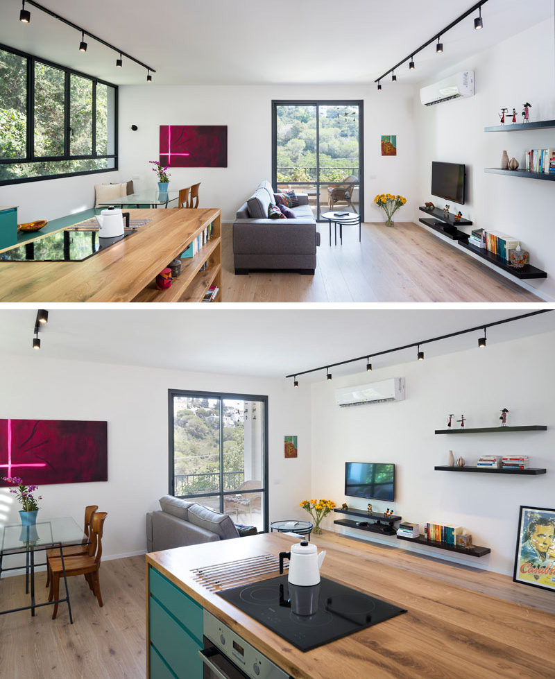 This open plan small modern apartment has a wood island with built-in book shelves the defines the kitchen area, while a gray couch sits in front of the TV wall, which has black track lighting and floating black shelves to allow for extra ornamental displays. 