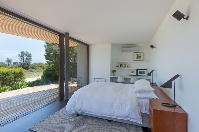 This modern master bedroom is lightly decorated with a small built-in desk and floating shelves above, as well as a wood headboard. The bedroom with dark wood flooring, opens up to a wood patio with views of the surrounding landscape.