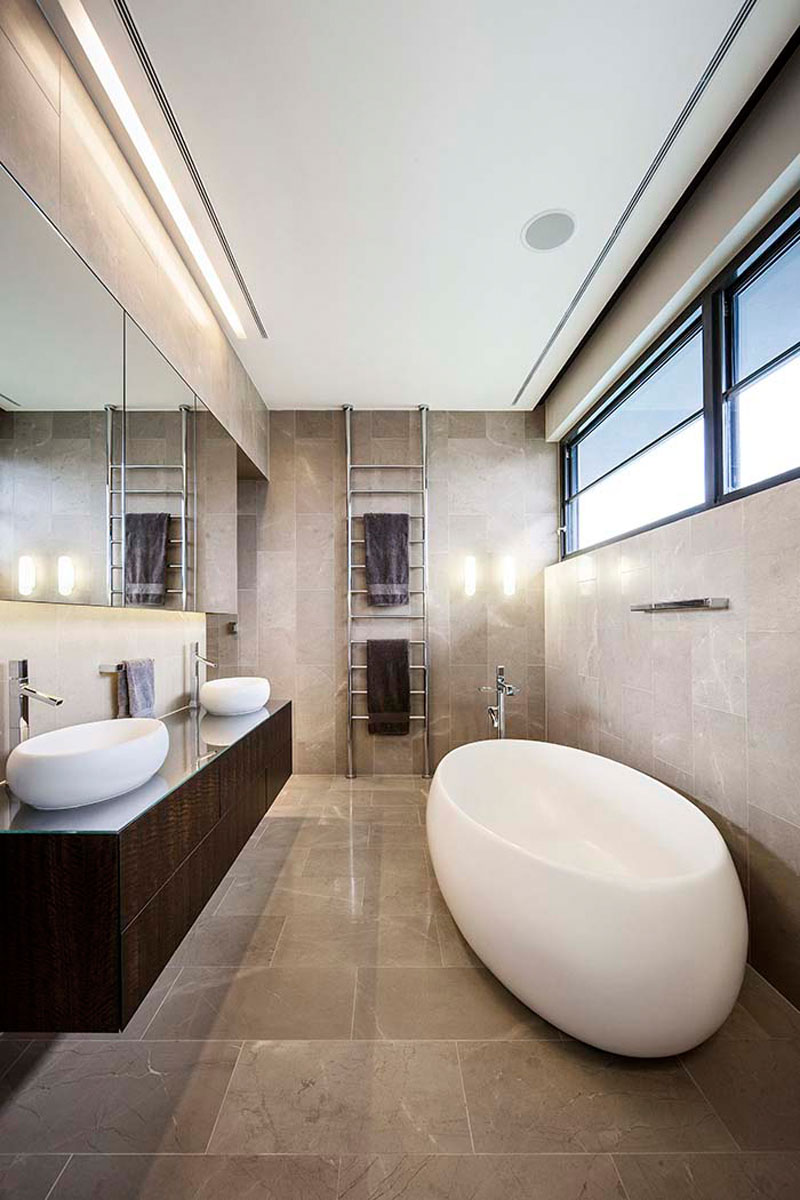 In this modern light grey tiled bathroom, a large rectangular mirror reflects light from the hidden lighting in the ceiling, and from the windows above the freestanding white bathtub.