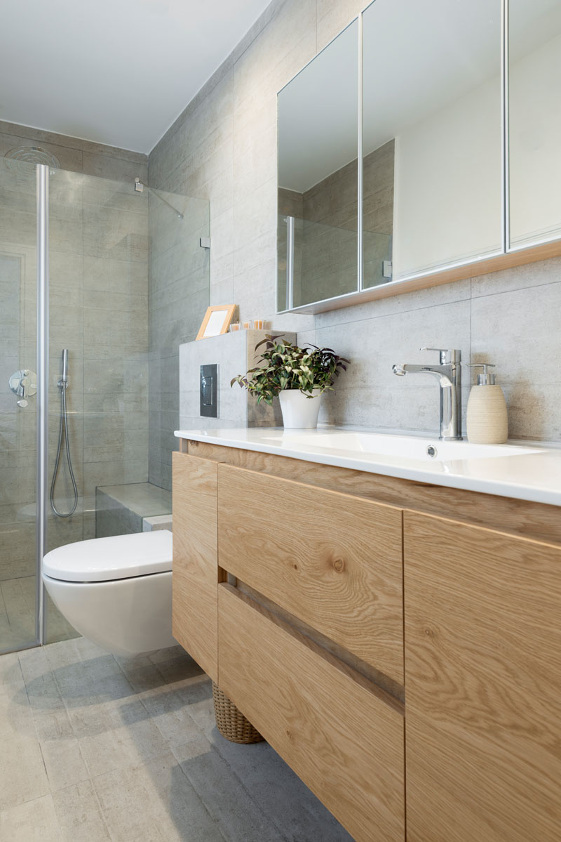 In this modern ensuite bathroom there is a glass surround shower with light colored tile. The wood cabinets and drawers below the white sink provide plenty of storage for toiletries.