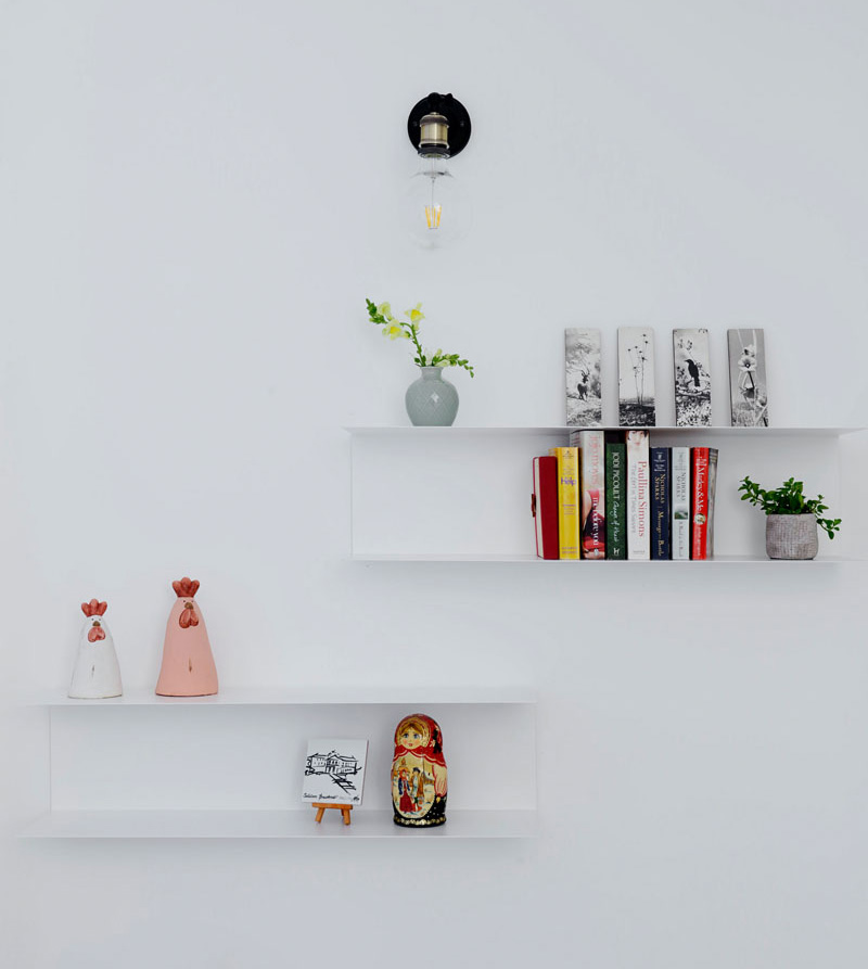 In this modern apartment, a set of minimalist white floating shelves are mounted below a single bulb, black wall sconce. The ornaments and books displayed add a pop of color to the white wall.