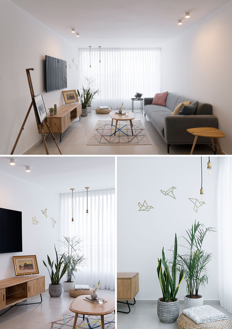 In this modern apartment living room, opaque white curtains cover a floor-to-ceiling window. A grey upholstered sofa, and light wood furniture give the room a warm feel. Two delicate pendant lights hang in front of gold origami bird wall decals.