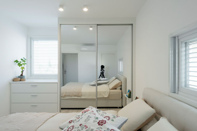 In this modern master bedroom, the decor is almost entirely white making it nice and bright. Placed beside a white built-in dresser is a closet with a sliding mirrored door that helps to reflect the light.