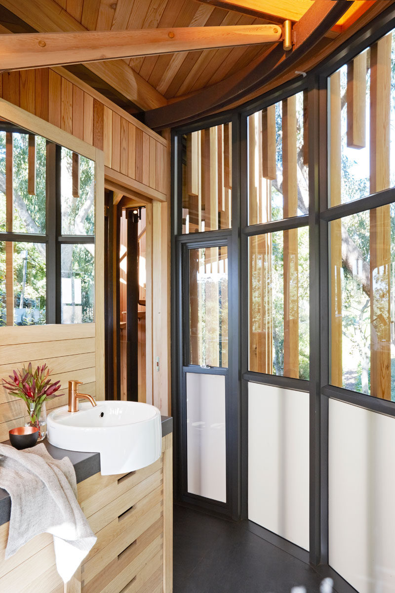 Paneled black framed windows compliment the dark tiled floor in this bathroom, while a white circular sink with a brass faucet rests above the light wood vanity.