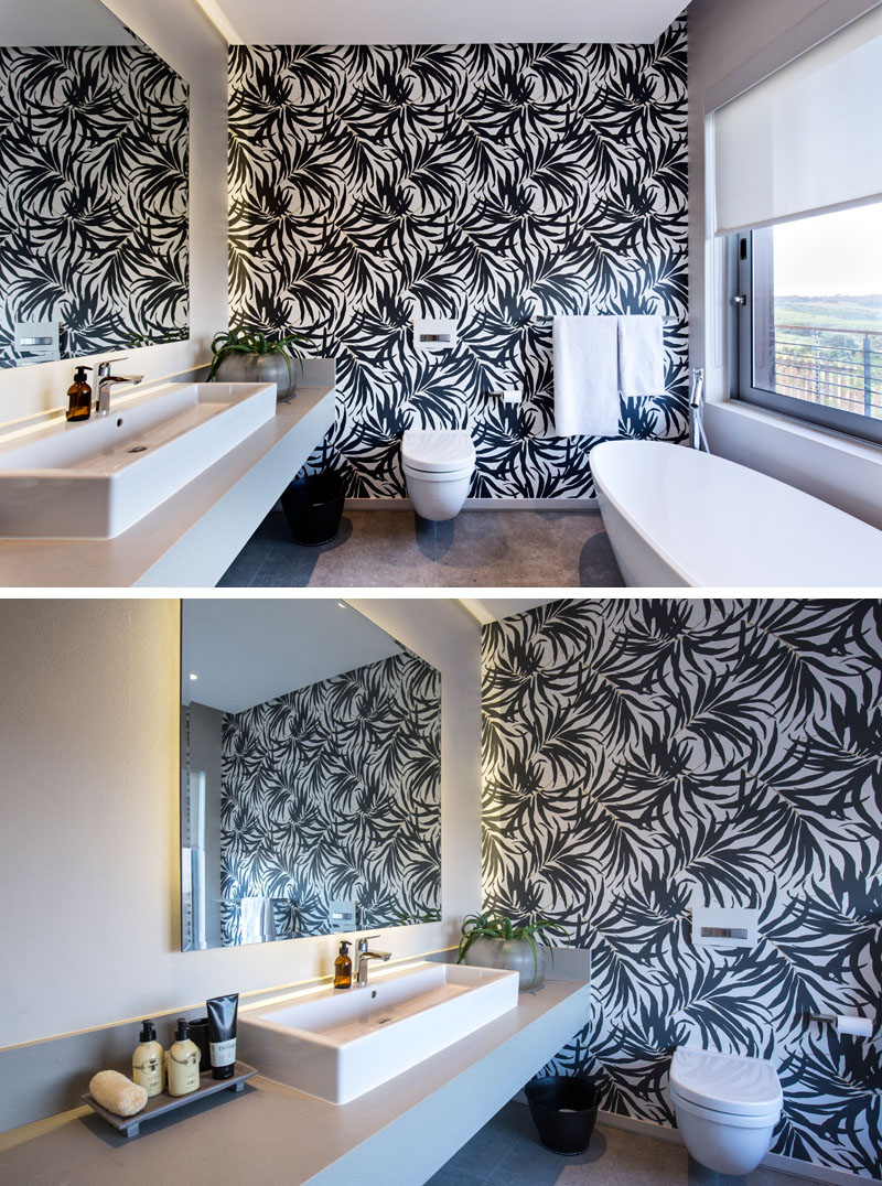 The black and white wallpaper on the accent wall of this modern bathroom is bold, making the white bath, toilet, vanity and decor standout. A large window with a white pull down privacy blind, provides views of outside when wanted.