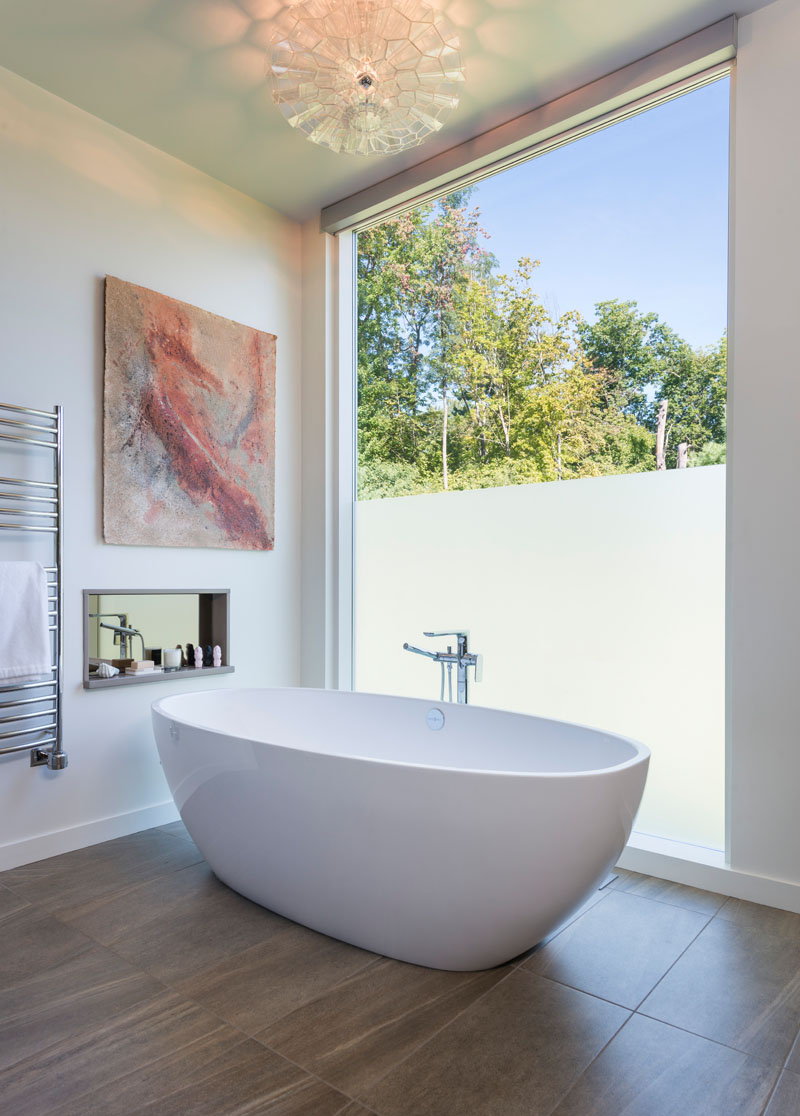 In this modern bathroom, an oval freestanding bathtub sits in front of a partially frosted floor-to-ceiling window.